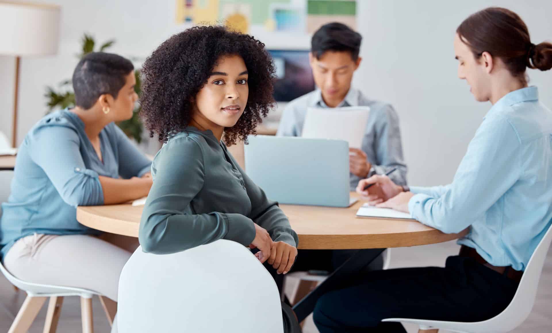 Frau blickt in die Kamera, während ein Team von Fachleuten im Hintergrund an einem Meeting arbeitet.