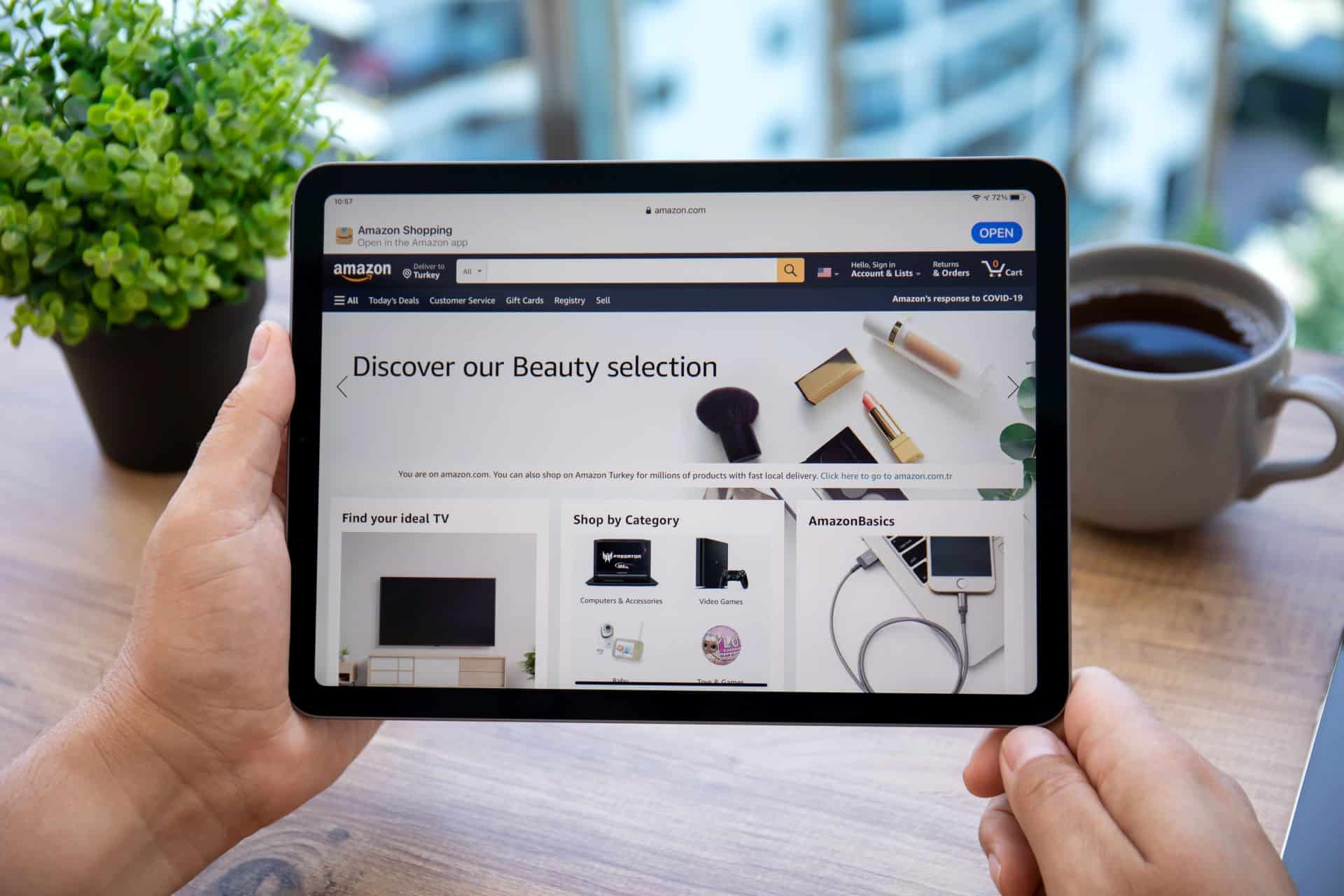 Person hält ein Tablet mit der geöffneten Amazon-Webseite, auf der Beauty-Produkte und Kategorien wie TV, Elektronik und AmazonBasics angezeigt werden. Im Hintergrund sind eine Pflanze und eine Tasse Kaffee zu sehen.