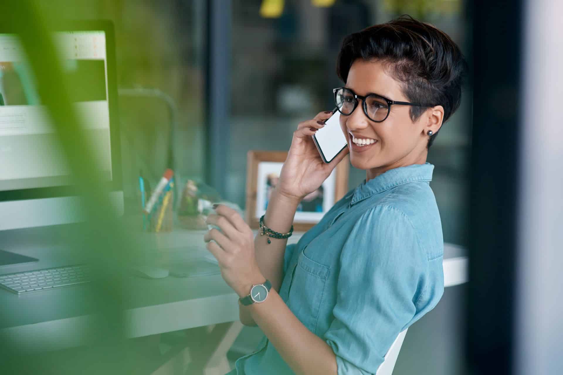 Eine lächelnde Frau mit Kurzhaarschnitt und Brille telefoniert in einem modernen Büro. Sie sitzt an einem Schreibtisch vor einem Computerbildschirm, während im Hintergrund Büromaterial und ein eingerahmtes Foto zu sehen sind.