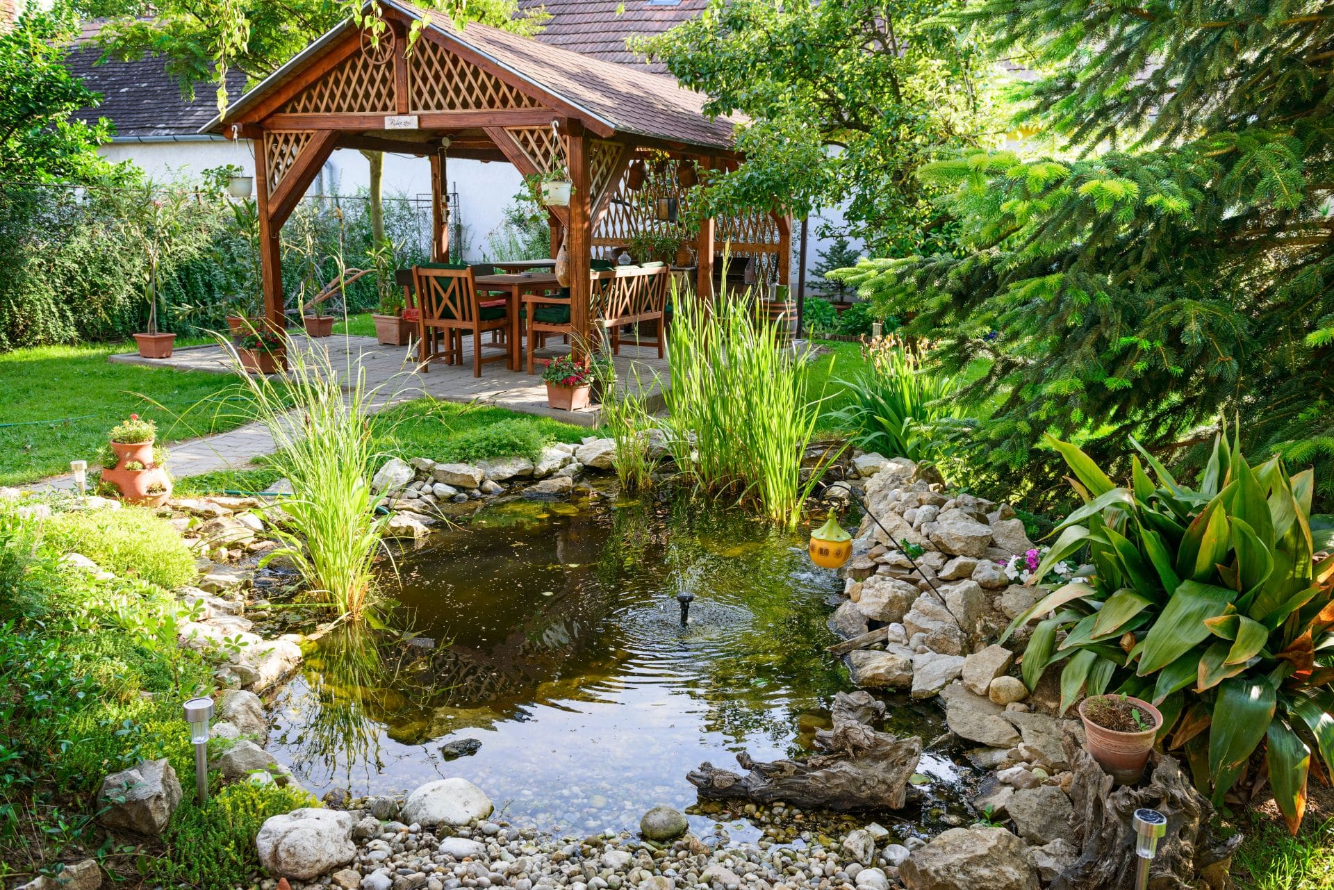 Ein wunderschöner Garten mit Teich und einem gemütlichen Sitzbereich unter einem hölzernen Faltpavillon, umgeben von grünen Pflanzen.