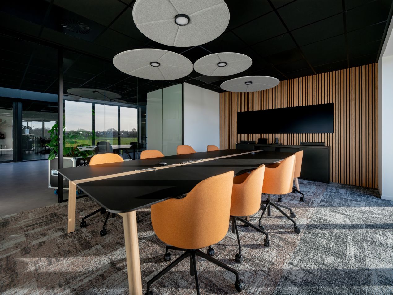Moderner Meetingraum mit runden Akustikdecken-Elementen und vertikalem Wandpaneel aus Holz hinter dem Bildschirm, kombiniert mit ergonomischen Stuehlen